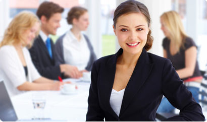 plisk-home-agents Photo of smiling conservatively dressed business woman facing the camera, standing in front of a group of other co-workers who appear to be meeting.